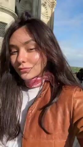 The video, likely a selfie video, shows a young woman with long, wavy, dark brown hair standing on a balcony or terrace of a castle or historical building. She is wearing a tan leather jacket and a patterned scarf around her neck. In the background, there are other people, possibly tourists, and a view of the surrounding landscape. The building itself is made of light-colored stone, with details like towers and decorative elements visible. The sky is blue with some clouds. The woman appears to be enjoying the view and the castle's ambiance.