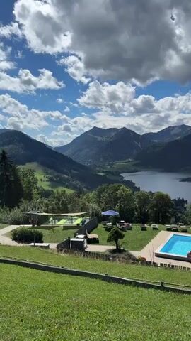 The video pans from left to right across a scenic mountain landscape. In the foreground, a grassy hill slopes down towards a swimming pool. Several people are visible in and around the pool, with at least two individuals swimming. Beyond the pool, there is a seating area with chairs and tables, some with umbrellas. A small building with a pink roof is also present. The middle ground features a dense forest and a body of water, likely a lake, with a small island. In the background, majestic mountains rise against a bright blue sky dotted with white clouds. The overall scene depicts a serene and picturesque vacation setting.