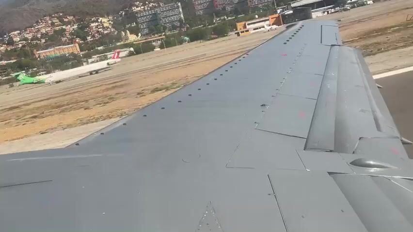 From the perspective of an airplane window, the plane's wing is visible, along with other aircraft on the tarmac.  The aircraft have various color schemes and some have names, such as CONVIASA, printed on the fuselage. In the background is a city with buildings and hills. The sky is slightly hazy. The airplane is moving.