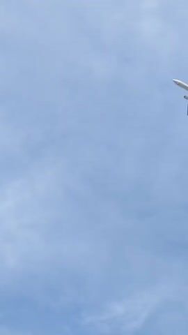 The video shows an aircraft flying in the sky. The sky is light blue with a cloudy white overcast. The aircraft is white with green accents, and the logo on the fuselage appears to read "SAUDIA."  The plane moves from the upper right corner towards the upper center of the frame, suggesting it's flying either away from the camera or directly overhead. The perspective is from ground level, looking upwards.