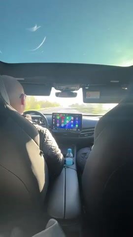 The video shows the interior of a car while it is being driven on a road. The camera is positioned behind the two front seats, looking forward towards the driver and the dashboard. The driver, a bald man wearing glasses and a dark jacket, is visible at the steering wheel. The car has a large, panoramic sunroof which allows a view of the bright, sunny sky above. The dashboard features a large touchscreen display with various icons. A water bottle is visible in the center console. The car appears to be driving on a relatively straight road with fields visible in the distance. The camera angle changes slightly throughout the video, sometimes focusing more on the road ahead and sometimes on the sunroof.