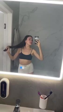 A young woman with long dark hair takes a selfie in a well-lit bathroom mirror. She is wearing a black bralette and white shorts. Her left arm is bent at the elbow and her hand is near her head, with her hair extending over her shoulder. Her right arm is extended, holding a smartphone to take the photo. The bathroom features marble-patterned walls, a white countertop, and various bathroom accessories visible in the reflection, including toothbrushes in a holder. A glowing light surrounds the mirror, illuminating the scene.