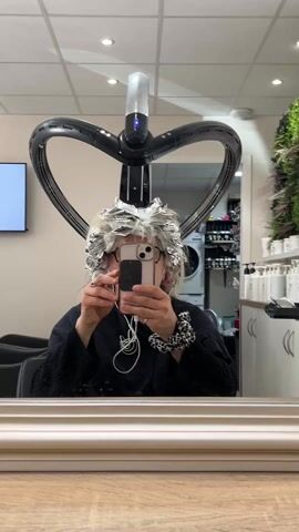 A woman sits in a salon chair in front of a mirror, holding up her phone to take a selfie. She is wearing glasses and has foil in her hair, presumably for a hair coloring or highlighting process. Above her is a large, modern hair dryer, which has a circular design with three arms.  In the background, there is a television mounted on the wall, showing an image of a building. A washing machine is visible in a doorway. To the right, a counter holds an assortment of hair care products, and behind it is a decorative plant wall.  The salon appears bright and clean with a contemporary design. The woman seems to be passing the time by taking a picture while waiting for her hair treatment to process.