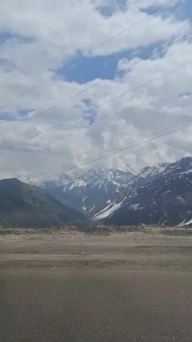 The video shows a mountain range with snow-capped peaks under a partly cloudy sky. The foreground consists of a flat, barren, rocky area. Overhead power lines are visible. Music is playing in the background.