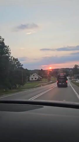 The video shows a sunset over a rural landscape, viewed from inside a vehicle. The sun is visible low in the sky, casting a warm glow and creating lens flares. A road with double white lines runs through the scene, with a barrier on the left. On the left side of the road, there are houses and trees. On the right side of the road, a black van with text on the back is visible, followed by a red car. The sky is filled with clouds in shades of pink, orange, and blue.