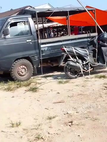 A black pickup truck with an orange tarp covering the bed is parked on a dirt lot. A black motorcycle is tied to the side of the truck. There are several people visible in the background, some of whom appear to be vendors at a market. The environment appears to be a rural or outdoor market setting.