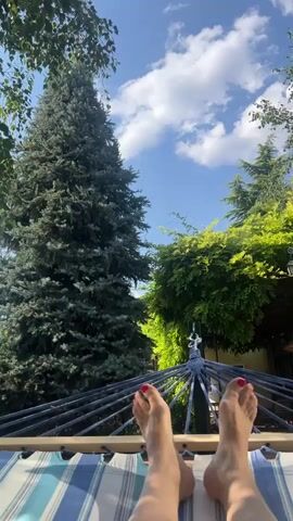 The video shows a first-person view of someone lying in a hammock outdoors on a sunny day. The camera is pointed upwards, showing the person's feet and legs in the foreground. The hammock is striped with blue and white. In the background, there are trees and a bright blue sky with white clouds. A large, dark green evergreen tree is on the left, and a lush green tree with lighter green leaves is on the right.