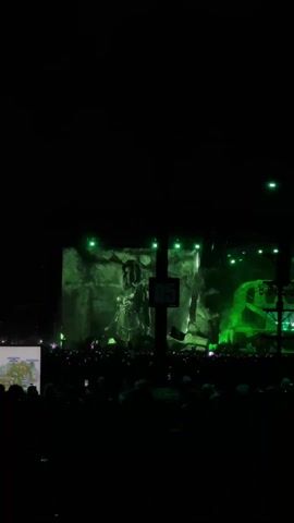 The video captures a large, night-time outdoor music concert or festival in Indio, United States of America. A vast crowd of people is visible, facing a brightly lit stage in the distance. The stage is predominantly illuminated with vibrant green lights, creating an ethereal and energetic atmosphere. A large screen on the stage displays various green-toned visuals, including what appears to be a human figure or face and abstract rock-like formations, changing throughout the clip.

In the foreground, a tall, dark pole with multiple lighting fixtures stands prominently, bearing a clear "D5" sign. A Mexican flag is briefly visible on this pole. Below the "D5" sign, the text "KEEP OFF" is visible on another small sign. To the far left, a screen or board displays a detailed map of the event grounds. The crowd appears to be engaged, with many holding up their phones, likely recording or taking photos. The overall scene suggests a high-energy musical performance at a large-scale event.