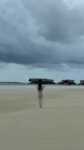 A wide shot depicts a woman walking away from the camera on a beach towards buildings on stilts in the water. The woman has dark hair, is wearing a swimsuit and a sheer white garment, and she has her hands up in the air. The beach is sandy and the sky is overcast with dark, heavy clouds. There is a slight breeze visible in the way the woman's garment moves.