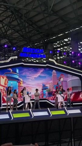 A group of nine young people, appearing to be performers, are on a stage. The stage is set up with a large, colorful LED screen in the background displaying a cartoonish urban landscape with a bright sky. Above the screen, a neon sign reads "MADOK HAWAII". The performers are arranged across the stage, some facing the audience, some facing each other, and some in profile. They are dressed in casual attire, including t-shirts, shorts, and skirts. One performer in the center is wearing a white outfit that appears to be a costume. Most of the performers are facing the front of the stage and seem to be engaged in a synchronized dance or presentation. The environment appears to be an indoor venue with a high ceiling and exposed industrial lighting. The overall impression is that of a performance or event.