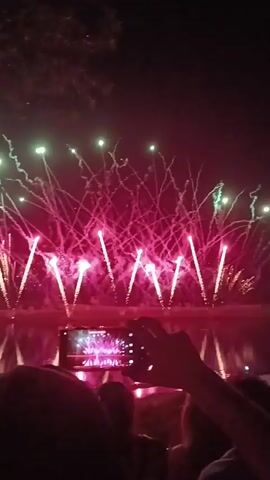 The video shows a nighttime fireworks display over a body of water, likely a lake or river, as evidenced by the reflections of the fireworks on the surface. In the foreground, the silhouettes of several people are visible, suggesting they are gathered to watch the event. One person in the center is holding up a smartphone horizontally, recording the fireworks. The phone screen is illuminated and displays a captured image of the fireworks. The fireworks themselves are colorful, with bursts of pink and green light, and trails of sparks ascending into the dark sky. The overall scene conveys a festive atmosphere, with people observing and documenting a celebratory event.
