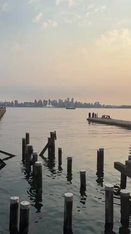 The video shows a view of a city skyline across a body of water. In the foreground, there are several wooden pilings in the water, some of which are broken or leaning. There is also a dock or pier extending into the water on the right side of the frame, with several people standing on it. In the middle ground, a ferry is moving across the water towards the left side of the frame. The city skyline is in the background, with numerous tall buildings. The sky is overcast with some clouds. The lighting suggests it is either early morning or late afternoon.