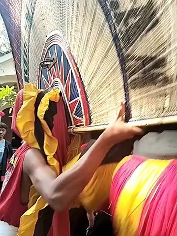 The video shows what appears to be a cultural procession in a neighborhood. The main focus is on an individual carrying a large, circular, and intricate piece, possibly part of a costume or decoration. It's made of light-colored material and trimmed with darker colors. The person carrying it is wearing a red and yellow outfit. Behind them, a group of musicians play instruments that look like angklungs, a traditional Indonesian instrument. A crowd of spectators, mainly young people and some adults, watch the procession from the side of the road. Houses and trees are visible in the background, suggesting the event is taking place in a residential area.