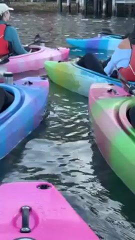A group of people are in colorful kayaks on the water. The kayaks are in pink, blue, green and purple colors. The water is choppy. The people are wearing life jackets. There are wooden pilings visible in the background.