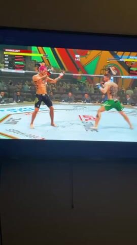 The video shows a person playing a UFC fighting video game on a TV. Two characters are facing each other in the ring. The character on the left has pink hair, black shorts, and tattoos. The character on the right has brown hair, green shorts, and tattoos. The character on the right throws a kick, and the character on the left dodges it. The game interface shows the names of the characters, their health bars, and the timer for the round. The background of the ring shows spectators and advertisements. The wall behind the TV is a light color, and there is a hanging cord visible.