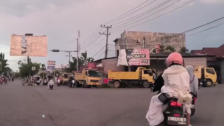 The video shows a street scene in Indonesia. Several yellow dump trucks are parked on the side of the road. Motorbikes are driving down the street. One rider is wearing a pink helmet and a white outfit. Another is wearing a hijab. There are several advertising signs visible. One sign is for the fabric brand Wadimor. Another sign is for mangoes. The sky is overcast.