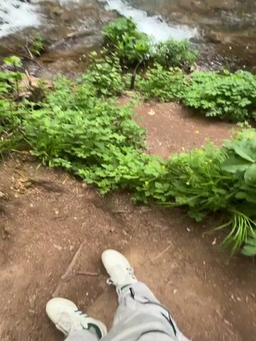 The video shows a waterfall cascading down a series of rocky ledges in a green forest. The camera starts low, showing the feet of a person standing on a dirt path surrounded by lush green plants, then pans up to reveal the waterfall and the surrounding forest. The water is clear and flows rapidly over the rocks. The forest is dense with green trees, creating a peaceful and natural setting.