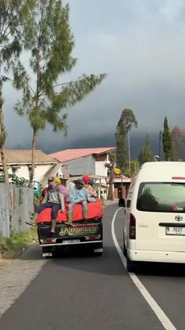 A black pickup truck drives down a paved road. On the truck bed, which is covered with a red tarp, sit five men. They are all wearing casual clothes and some have on boots. The truck has a license plate that says N 9279 TT. Next to the pickup truck, a white van drives in the same direction. In the background, there are houses, trees, and a dark, cloudy sky. The scene appears to be in a rural or mountainous area.