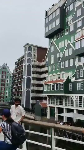 A video shows a picturesque canal-side scene with buildings designed in a traditional Dutch architectural style. The buildings are characterized by their steep gabled roofs, prominent white trim, and vibrant green facades, resembling stacked houses. The architecture is somewhat repetitive, with similar designs appearing across several structures. In the foreground, a wooden bridge with white railings spans the canal. Several people are visible on the bridge, some posing for photos and interacting. One man in a white patterned shirt stands near the railing on the left. Further along the bridge, a woman in a pink dress is looking to her left, and another woman in a black t-shirt and denim shorts is striking a pose, with a man in a black tank top holding a phone, likely taking a picture. Another woman in a brown shirt is walking away from the camera. The sky is overcast, suggesting a cloudy day. The water in the canal is calm. There are also glimpses of cafes and shops at the base of the buildings. The overall impression is a lively and aesthetically pleasing urban environment.