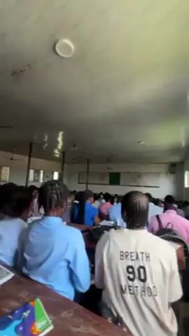 The video shows a crowded classroom from the back, with numerous students seated at desks. Most students are wearing light-colored tops, including blue, white, and pink. A teacher stands near the front of the room in front of a whiteboard filled with writing. Several ceiling fans are visible. The classroom has large windows on one side, providing natural light. The ceiling is low and painted white.