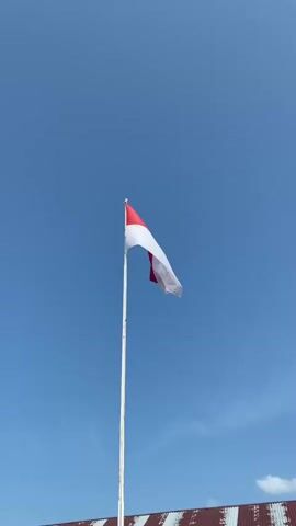 The video shows a close-up, low-angle shot of the Indonesian flag flying against a clear blue sky. The flag is white on the bottom and red on top, and it is attached to a white flagpole. The flag is billowing in the wind, and its movement is captured in slow motion. The bottom of the frame shows a blurred glimpse of a building with a red and white striped roof. The sky is a bright, cloudless blue, suggesting a sunny day. There are no people visible in the video.