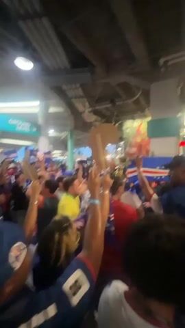 A crowd of people are gathered in an indoor area. Many are holding up flags and other objects. The flags appear to be of various countries, including what looks like the flag of Cape Verde. Some individuals are wearing blue shirts, and others are wearing yellow. The environment appears to be some kind of concourse or walkway, with a sign that reads "GROUND LEVEL" visible in the background. The atmosphere seems to be festive and energetic, with people cheering and waving.