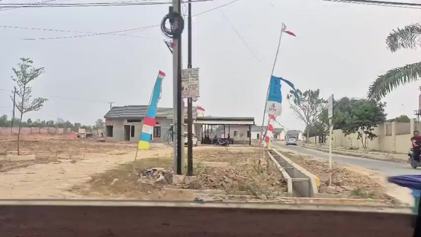 The video shows a developing real estate area with several unfinished houses and signs advertising a residential project. The environment is a dusty, open field with sparse trees and some construction materials scattered around. Red and white flags are prominently displayed, likely indicating national or project branding. A few vehicles, including a car and a motorcycle with a passenger, are visible on a dirt road leading into the property. In the foreground, the edge of a vehicle's interior is visible, suggesting the footage is captured from inside a car or van. The overall impression is of a new housing development in its early stages of construction, set in an undeveloped, possibly rural or suburban, landscape.