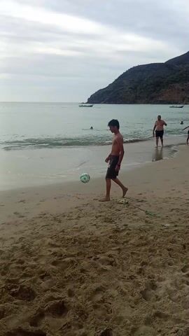 The video shows a beach scene with a group of people. The focus is on a young man who is juggling a soccer ball with his feet and then with his head. He is wearing dark shorts and is shirtless. There are several other people walking and standing along the shoreline and in the water. A few small boats are visible in the distance on the water. A hill covered with vegetation is visible in the background. The weather appears overcast.