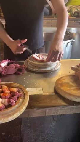 A person in a black shirt and apron is using scissors to cut up a cooked octopus on a metal counter. The octopus is large and bright pink, with visible suckers on its tentacles. Several wooden platters are on the counter, some containing pieces of octopus. In the background, there are stacks of wooden plates or coasters, and a large metal pot. The environment appears to be a kitchen or food preparation area. Another person's hand is visible in the foreground, holding a piece of what looks like food. The overall scene suggests the preparation of a seafood dish.