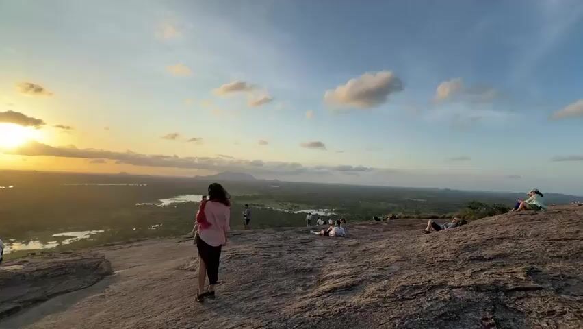 The video shows a group of people on top of a rocky hill, enjoying the sunset. The sun is setting over a landscape of trees, fields, and bodies of water. A few clouds are scattered in the sky. Some people are sitting on the rocks, talking and taking pictures. A woman in a pink shirt and black skirt is standing and looking at the view. The camera pans slightly to the left, showing more of the surrounding landscape and the people on the hill. The scene is peaceful and scenic.