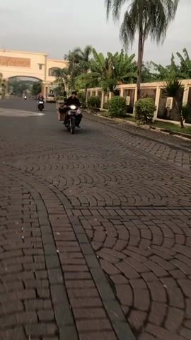 The video shows a paved road with a distinctive brick pattern. A person is riding a motorcycle with a box attached to the back, heading towards the viewer. In the background, a large building with an archway is visible, possibly an entrance. There are palm trees and other vegetation along the side of the road, enclosed by a wall. Another motorcycle is further in the distance, moving away from the viewer. The lighting suggests it might be early morning or late afternoon.