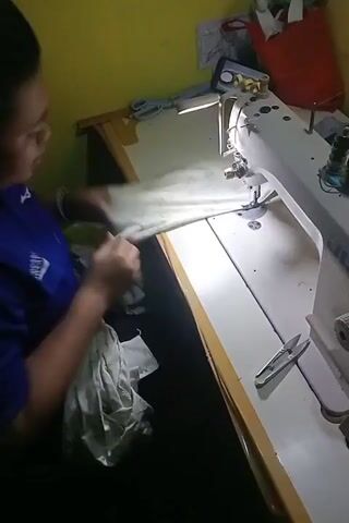 The video shows a person sewing on a white sewing machine. The person is wearing a blue shirt and is using their hands to guide the fabric. There are scissors on the table next to the sewing machine. The background is a yellow wall. The lighting is dim, with a bright light focused on the sewing machine.