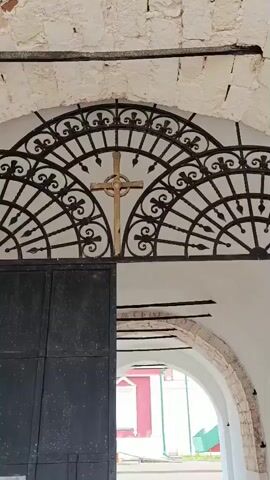 The video shows a view from inside an arched entryway. The entryway is constructed of white plastered stone with rough-hewn stone blocks visible along the archways. Above the arched opening, there is a decorative black wrought iron grate with a golden cross mounted in the center. The floor inside the entryway is paved with dark gray cobblestones.

The camera moves forward through the arched entryway, revealing a courtyard beyond. In the courtyard, a person dressed in dark clothing is visible, appearing to be walking away from the camera. Behind the person is a building with a prominent red facade, white trim, and a white-capped window. To the left of the red building, a green structure is partially visible. The ground in the courtyard appears to be paved with lighter-colored cobblestones or concrete. The lighting suggests it is daytime.