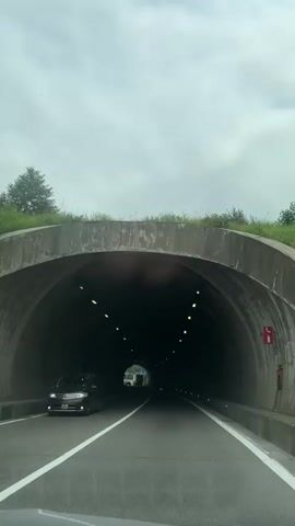 The video shows a view from inside a vehicle driving through a tunnel. The tunnel is concrete with lighting fixtures along the ceiling. There are other vehicles visible in the tunnel ahead, and the road is marked with white lines. The video appears to be shot on a cloudy day, as indicated by the overcast sky visible at the entrance of the tunnel.