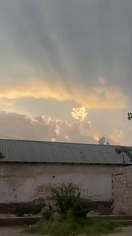 The video shows a low-angle view of a building's corrugated metal roof against a cloudy sky during what appears to be sunset. The sky is filled with dramatic clouds, with shafts of sunlight breaking through, creating a warm, golden glow. On the roof, a satellite dish is visible. The wall of the building beneath the roof is rough and shows signs of wear. Sparse vegetation grows in front of the wall and on the ground. The scene is static, with no discernible actions or interactions taking place.