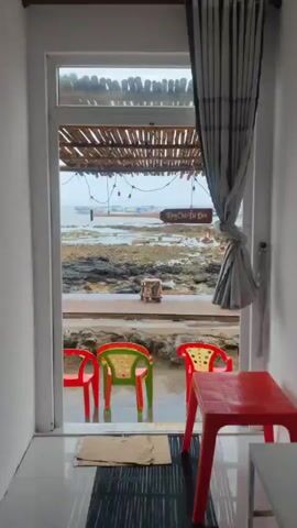 The video captures a view from indoors looking out onto a wet, rocky coastline under an overcast sky. Inside the room, a partial red plastic table is visible on the right, and a grey curtain is tied back beside an open doorway. Just outside the doorway, on a damp surface, sit three plastic chairs: two red and one green with red and yellow star patterns. A doormat lies on the floor inside.

Beyond the chairs, a wooden deck or walkway extends towards the water. The ground is a rugged expanse of dark rocks, suggesting a low-tide shore. A rustic canopy made of bamboo or thatch covers the deck area, with string lights strung underneath.

In the mid-distance, the ocean is visible, dotted with numerous small, brightly colored boats. A paddle-shaped wooden sign hangs from the canopy, displaying the Vietnamese text "Làng Chài Đá Đen" (Black Rock Fishing Village). Another small sign, partially visible on a post near the deck, reads "Cầu trường LÀ 300 M" (Bridge is 300m). The sound of water, possibly rain or dripping, is audible throughout the clip.