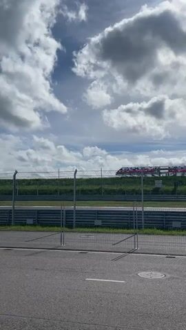 This is a vertical, first-person perspective video taken from a paved area next to a race track. The camera is looking towards a wire fence that separates the paved area from the grass and the race track beyond. Several cars are seen driving on the race track. The sky is partly cloudy with a mix of blue sky and white clouds. There are tall light poles visible on the side of the race track. The paved area in the foreground appears to be a parking lot or pit lane, with white painted lines and a circular manhole cover visible on the ground. The video captures the movement of the cars on the track and the general atmosphere of a race event on a cloudy day.