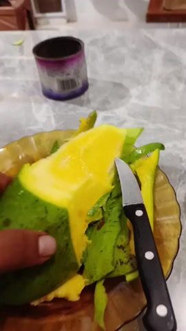 A person's hand holds a large, partially peeled mango over a brown, translucent plate. The mango has a vibrant green skin, and its exposed flesh is a rich, bright yellow, indicating ripeness. Several green mango peels are scattered on the plate, alongside a small knife with a black handle and a silver blade. In the blurred background, a metal can, possibly purple or blue with a white label and a barcode, sits on a light-colored, possibly marbled, table surface. The scene appears to be an indoor setting, likely a kitchen or dining area, where someone is preparing the fruit.