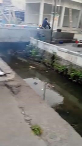 A man on a motorcycle rides past an open drainage ditch filled with murky water and debris. The ditch is lined with concrete and overgrown with plants. In the background, a building under construction is visible, with scaffolding and building materials scattered around. The scene appears to be an urban or semi-urban environment.