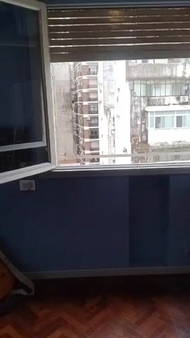The video begins with a view from inside a room, looking out a window with closed blinds. The window is then opened, revealing a view of several tall buildings. The camera angle tilts downward to show a flat rooftop area below, followed by a view of a small, overgrown garden area. The garden includes greenery covering walls, a small lawn, and a bare tree in the center. The video ends with the full view of the garden.