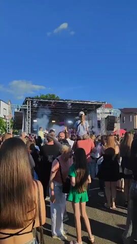 The video shows an outdoor concert with a stage set up under a clear blue sky. A crowd of people is gathered in front of the stage, many of them facing the performers. Smoke or fog is being emitted from the stage, illuminated by purple lights. A young girl with blonde hair is sitting on someone's shoulders, facing the stage and appearing to enjoy the music. Other individuals are visible in the crowd, some with their phones out, possibly recording the event. The overall atmosphere appears to be festive and lively.