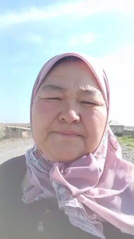 This is a selfie video featuring a woman wearing a pink hijab and a dark top. She is looking directly at the camera with a neutral expression. The setting appears to be a rural area with a gravel road in the foreground. In the background, there are several simple buildings, fences, and utility poles under a clear blue sky. The lighting suggests it is daytime, likely late morning or early afternoon, with bright sunlight. The overall atmosphere is calm and peaceful, with no discernible activity other than the woman recording herself. There is no visible text in the media file. Based on the provided location context, this scene is set in Chon-Kara, Kyrgyzstan.