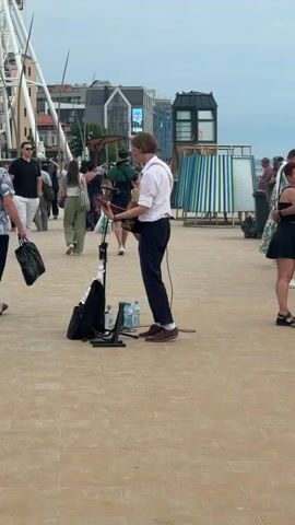 A street performer is playing an acoustic guitar on a paved walkway with a beach and the ocean in the background. The performer is wearing a white shirt and dark pants. There are several people walking in the background, some of whom are looking at the performer. A Ferris wheel is visible in the far left of the frame. The sky is overcast.