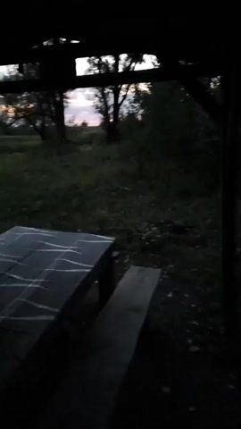 The video shows a view from under a roofed structure, looking out at a field at dusk. In the foreground, there is a picnic table and a bench, partially obscured by shadows. The table has white markings on its surface, possibly paint or chalk. Beyond the structure, a grassy field stretches towards a line of trees. The sky above the field displays the soft colors of sunset, with scattered clouds. The overall lighting suggests it is either early morning or late evening, with the sun low on the horizon. There are no people visible in the video.
