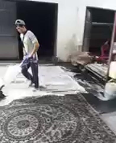 The video shows a person cleaning a large, patterned rug with a floor buffer. The person, wearing a light-colored shirt, dark pants, and a cap, is operating the buffer which is covered in foam. The rug is laid out on a light-colored surface, possibly concrete, with some debris visible around the edges. In the background, there is a large, dark opening that resembles a garage door or an industrial entrance, with a lighter-colored wall to the left. The scene appears to be outdoors or in a partially enclosed space, given the lighting and the open background. The activity suggests a professional or domestic rug cleaning process. No text is visible in the video. The lighting suggests it could be daytime, but the exact time of day and weather conditions are not discernible. The setting appears to be a somewhat utilitarian or industrial area, possibly a workshop or a yard.