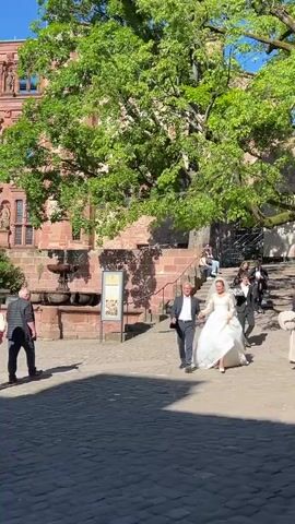 The video captures a joyful wedding couple, with the bride in a flowing white gown and the groom in a dark suit, walking hand-in-hand across a sunlit cobblestone courtyard. They appear to be smiling and happy. Surrounding them are several other individuals, some walking casually, others observing.

The setting is an outdoor plaza bordered by magnificent historic buildings constructed from reddish stone or brick, featuring multiple stories, ornate windows, and intricate architectural details. Large, lush green trees provide shade and frame the scene. To the left, a stone fountain is visible. The sky is a clear, bright blue, suggesting a sunny day.

In the background, a large scaffolding structure draped with a banner displays the text "HEIDELBERGER FESTSPIELE." A yellow and green flag can be seen flying from a flagpole on one of the older buildings. This picturesque scene is set within the grounds of Heidelberg Castle in Heidelberg, Germany.