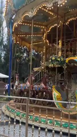 The video shows a carousel with horses and lights spinning in a circular motion. It's surrounded by a metal fence with decorative patterns. In the background, there are trees and a white structure with people visible. The carousel's ornate design features gold accents and multiple levels.