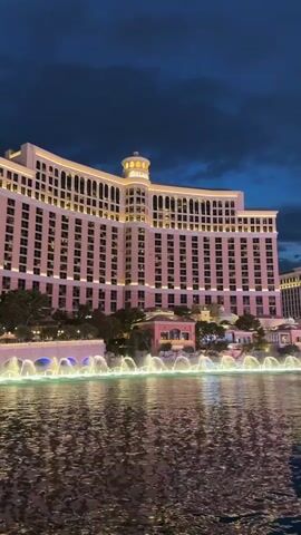 This is a static video of the Bellagio hotel and its fountains in Las Vegas at dusk. The hotel is a large, pink building with many windows, illuminated by exterior lights. In front of the hotel, a large body of water with illuminated fountains is visible. The sky above is a dark blue with clouds.