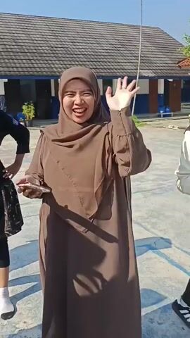 This is a video of a group of young women outdoors, seemingly on a basketball court. They are all wearing hijabs, which are Islamic headscarves. The women are smiling and interacting with the camera. One woman in the center is laughing with her mouth open and has her hands in a heart shape. Another woman on the right is waving and smiling. The woman on the far left is also waving. The background shows a building with a tiled roof and a basketball hoop. The sun is shining, casting shadows on the ground. The overall impression is one of a happy and informal gathering.