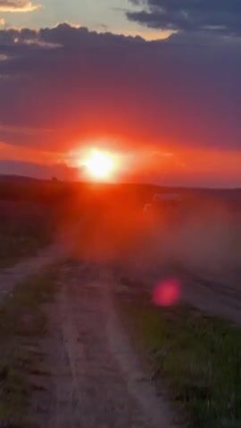 The video shows a dirt road stretching towards a bright sunset with orange and red skies. Some clouds are visible above. In the distance, a vehicle travels along the road, kicking up dust. There's grass on either side of the road. The overall scene is rural and captures the beauty of the sunset.